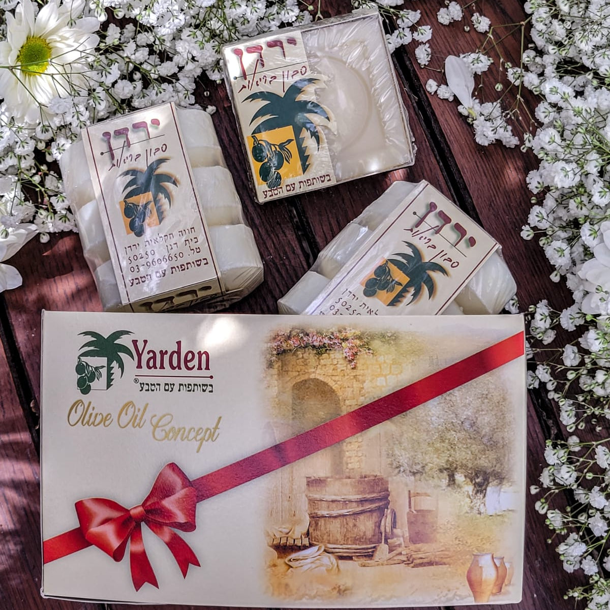 A gift box of Yarden olive oil soap with the packaging displaying an illustration of a palm tree and an olive press, accompanied by two individual soaps wrapped in decorative paper with a red ribbon on top, all presented on a bed of white flower petals.